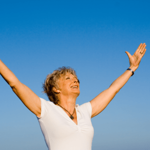 A woman with short gray hair with a white top holding her hands up in the air with the sky in the background