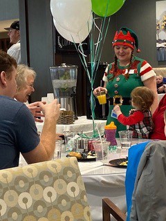 A family sitting around a table with White and Green balloons in a dining room. A baby is looking at a server holding orange juice and wearing an elf costume and the family is laughing.
