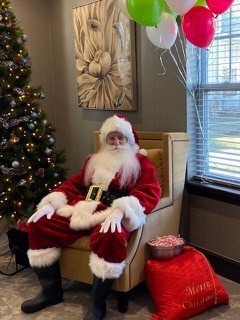 A man dressed as Santa sitting in a brown chair next to a decorated Christmas Tree and gift bag. Red, green, and white balloons behind him. Gray walls and brown carpet