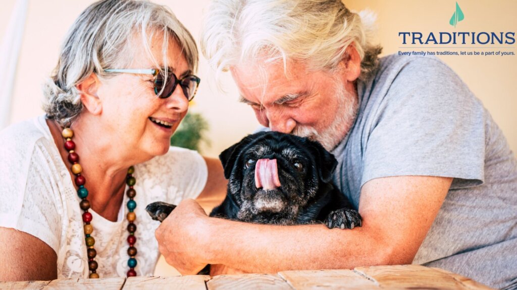 A senior man kissing a small black dog on the head and a senior woman sitting beside him smiling with the Traditions logo in the top right corner
