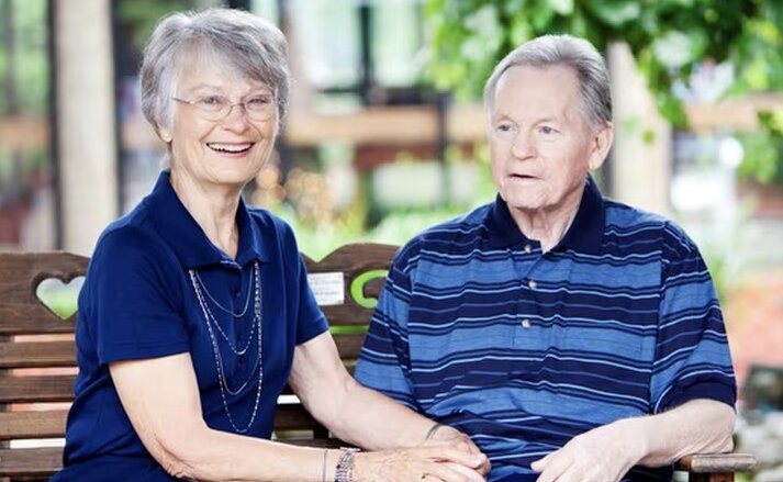 An elderly couple, in matching blue shirts, sit on a park bench together holding hands and enjoying the calm weather. Traditions at North Willow in Indianapolis, IN.